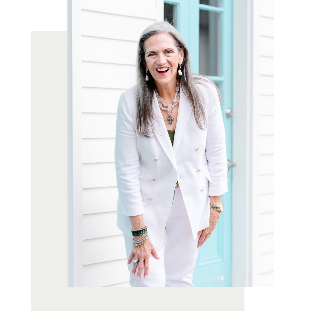 Smiling woman with long gray hair in a white blazer stands joyfully in front of a turquoise door. She wears layered necklaces and bracelets.